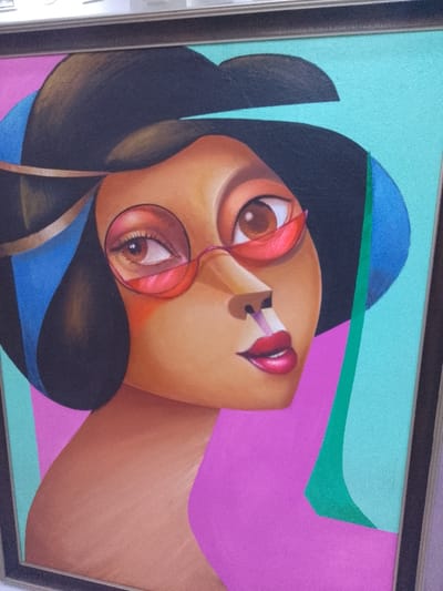A photograph taken in Ikate, Nigeria, displays a framed, stylized painting of a female subject. The artwork depicts a bust-up portrait with cubist-inspired elements, featuring a woman with warm brown skin, prominent brown eyes, and dark, short hair. She wears round, red-tinted spectacle lenses over her left eye, with a partial red frame extending near her right eye. Her lips are full and colored reddish-pink. The background is segmented into distinct color blocks: light teal in the upper right quadrant and vibrant magenta extending from the lower left. A thin, vertical green stripe separates the teal and magenta along the right side of the canvas. Dark blue geometric shapes are visible near the subject's head on both sides. The painting is enclosed within a dark frame. The photograph is captured at a slight upward angle.