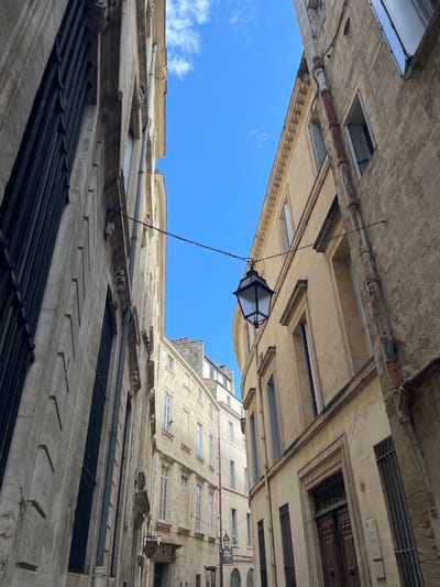 The image is a low-angle shot of an alleyway in Montpellier, France. The camera is tilted upwards, capturing the architectural details of the surrounding buildings and the blue sky above. The buildings are constructed from light-colored stone, with multiple stories and various window styles.  A black metal lantern hangs from a chain between the buildings.  Pipes and other architectural details are visible along the building facades. The sky is a clear blue with some scattered clouds.