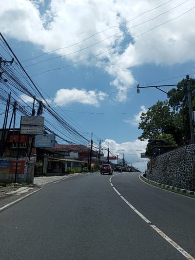 This image captures a daytime street scene, with the setting provided as the city of Bagochen Boolingpang Ueling, Bhutan. The perspective is from an asphalt road that gently curves to the right, under a clear blue sky interspersed with scattered white clouds.
On the left side of the road, a complex network of black overhead electrical and communication wires descends from utility poles. Several signs are affixed to these poles or adjacent structures. A white rectangular sign displays "NOTARIS" and further text in Indonesian, including "S. MARGARETTA SH", "PPAT", and "Jln." Below this, a blue rectangular sign features an arrow pointing right and the Indonesian text "MUSEUM ANGKASA PURA". Another partially visible red and white sign below reads "LITER PRAKTIS" and "TOKO SICI". Behind these, various buildings, some with red-tiled roofs, line the street, appearing to be commercial establishments or residences.
The dark gray asphalt road includes a faded white dashed line marking the center and a solid white line along its right edge. A red compact SUV is visible driving away from the viewer in the middle distance, with other vehicles discernible further down the road.
The right side of the road is defined by a low patterned stone retaining wall, composed of irregular dark and lighter gray stones. A light-colored concrete curb or barrier separates the road from this wall. Green tree foliage extends above the wall. A utility pole with a horizontal arm supports a street lamp projecting over the road.
Overhead, the bright blue sky with white clouds is traversed by several thin, dark parallel lines, consistent with power or communication cables. No individuals are explicitly visible.