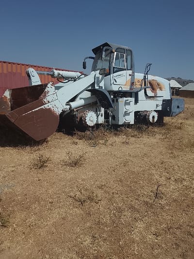 The scene, located in Bassa, Nigeria, displays an outdoor area under a clear, cloudless blue sky. A large, white and light blue YUTONG wheel loader, exhibiting considerable rust, faded paint, and peeling areas, occupies the center. Its prominent front bucket is significantly rust-colored (reddish-brown). The ground consists of dry, sparse, golden-brown grass and soil, with a few small green shrubs scattered throughout. To the left background, a reddish-orange shipping container is partially visible. In the right background, several small structures with light-colored roofs, possibly rural dwellings, are present, with distant hills visible beneath the horizon. No individuals or immediate activity are observed.