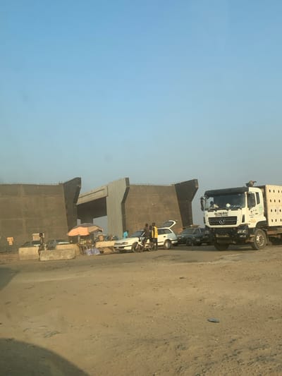 A street scene features several vehicles, including a large white truck and multiple cars parked along the roadside. People stand near one of the cars. Adjacent to the vehicles is what appears to be a concrete structure, possibly a bridge or building under construction. A vendor is positioned near the structure, with a small stall and a pink umbrella providing shade. The scene is bathed in natural light under a pale blue sky. The ground is dusty, suggesting an arid or construction-heavy environment.