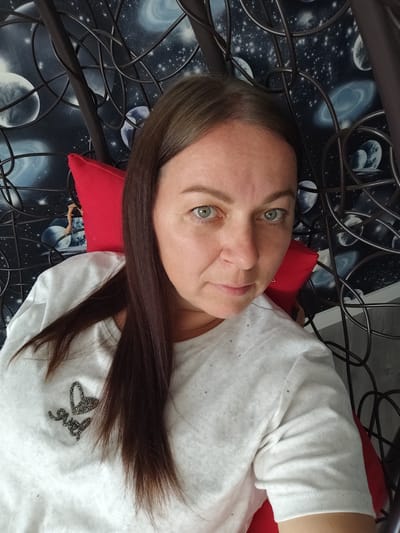 A woman with shoulder-length brown hair and green eyes is seated in a chair with a red cushion. She is wearing a white t-shirt with a beaded heart and the word "love" on it. The background is a dark wall with a space-themed wallpaper featuring planets and stars. The chair has a dark, intricate metal frame. The image appears to be taken indoors, likely in a residential setting, possibly in the city of Narach, Narachsky District, Minsk Region, Belarus.