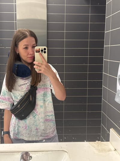 A woman takes a selfie in a public restroom. The background features black, rectangular tiles covering the wall. The woman wears a tie-dye t-shirt, a blue face mask around her neck, and a black fanny pack across her body. She holds a white smartphone with a visible camera in her right hand. The restroom includes a white sink and a paper towel dispenser above her head. It's difficult to confirm the location is specifically in Bol'shie Kabany, Russia, without additional information.