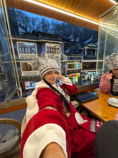 An individual is seated indoors at a wooden table, taking a selfie, and smiling at the camera. This person, of apparent European descent, is adorned in a red traditional Chinese-style robe with white trim, a fur-lined collar, and elaborate silver ornamentation, including a large, ornate silver headdress characteristic of Miao ethnic attire. Several dark braids with silver accents are visible over their left shoulder. A second individual, partially visible to the right, also wears a similar silver headdress and a pink puffy jacket, looking down. On the table between them rests a black folder, a smartphone, and a white dish with blue patterns.

The scene is set in Fenghuang Ancient Town, China, at dusk or night. Through a large window overlooking a river, traditional Chinese buildings with multi-tiered roofs and balconies are visible across the water. These buildings are extensively illuminated with white string lights and strip lighting along their edges and windows, creating a vibrant evening atmosphere. Distant hills with sparse trees are visible under a dark blue sky above the illuminated structures. The interior space features a warm, amber light strip running along the ceiling.
