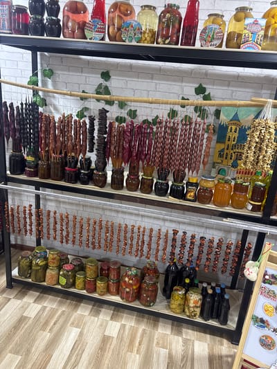 The image depicts an indoor retail setting, likely a market stall or shop, located in Manavi, Georgia. The scene is dominated by a multi-tiered display of various food products.
The background consists of a white wall with a faux brick pattern. The floor is covered with light-colored wooden laminate planks arranged in a staggered pattern.
The display includes:
1. **Top Shelf:** A black shelf holds a row of large glass jars. These jars contain preserved fruits and liquids, including dark jams, red fruit preserves, and whole light-colored fruits (possibly peaches or plums) in syrup. Small, colorful cardboard cutouts depicting stylized buildings are positioned between some jars.
2. **Mid-Level Hanging Rack and Shelf:** A wooden crossbeam is strung with numerous strings of *churchkhela*, a traditional Georgian confection made of nuts coated in thickened fruit juice. The *churchkhela* vary in color, ranging from dark red/purple to various shades of brown. Alongside these are strings of dried fruits or nuts, presenting a darker, more irregular texture. Below this hanging display, on a black shelf, are multiple smaller glass jars containing dark jams or honey, a jar of instant coffee labeled "Jacobs Monarch," and another labeled "Nescafe Gold." Several dark liquid-filled bottles are also present. A painting depicting a yellow building with a blue roof is affixed to the wall behind the hanging items. Artificial green leafy vines are draped over the wooden crossbeam.
3. **Lower-Level Hanging Rack:** A metal rack below the mid-level display features multiple strings of small, uniformly arranged dried fruit segments, light brown/orange in color, possibly dried apricots or other fruit pieces.
4. **Bottom Shelf:** A white shelf supports a long row of glass jars of various sizes. These jars primarily contain pickled vegetables, including cucumbers, mixed vegetables, and preserved tomatoes with garlic. Several dark-colored plastic bottles are also arrayed on this shelf.
5. **Right Side:** A portion of a wooden easel-like display board, featuring various magnets and small souvenirs, is visible on the far right. A small, light-colored, heart-shaped knitted ornament hangs from a string at the top right of this display.
No individuals are present in the scene. The items are static and arranged for retail presentation.