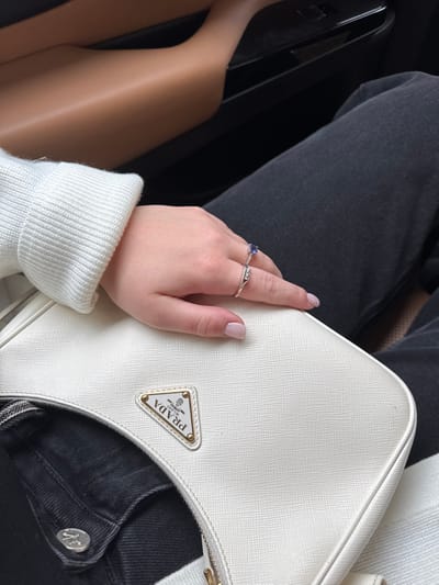 The image captures a close-up view from inside a vehicle, located in Dubai, United Arab Emirates. A person's left hand, displaying fair skin and light pink manicured fingernails, is centrally positioned. The hand wears two rings on the ring finger: an upper ring featuring an oval blue gemstone (likely sapphire) set in silver-toned metal, and a lower, narrower silver-toned band. The wrist is covered by a white ribbed knit sweater cuff. The hand rests on a white, textured Prada handbag, which prominently displays a gold-toned triangular Prada Milano logo. Below the handbag, a portion of dark gray or black denim jeans with a silver-toned button is visible. The background shows elements of the vehicle's interior, including a light brown or tan panel to the upper left and a darker panel with a visible control switch, likely for a window, below it.