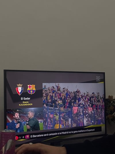 The image shows a television screen displaying a soccer game between FC Barcelona and El Sadar, broadcasted live on DAZN. The game's title card features the team logos and the words "El Sadar, Directo #LALIGAENDAZN." The screen also displays the score of the game "1 - 2". A crowd of spectators is seen in the background. A text overlay in Spanish reads, "El Barcelona será campeón si el Madrid no le gana mañana al Espanyol." A hand and a can of soda are visible in the foreground to the right of the television screen. The background consists of a plain white wall.