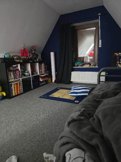 The image depicts a bedroom in Vreden, Germany, located at the coordinates 52.039 latitude, 6.849 longitude. The room features gray walls, a blue wall with a window and dark curtains, and carpet flooring. A large bookcase with multiple shelves stands against the left wall, filled with books and various items. A bed with a gray blanket and blue pillow sits on the right side of the room. A game mat is spread out on the floor in front of the bed. The bed is situated near a window with white blinds. The window is visible from outside. The room is illuminated by natural light from the window. No people are present.  The image appears to be taken from a low angle, facing the bed.