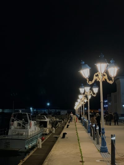 A nighttime scene features a row of ornate lampposts illuminating a concrete boardwalk along a harbor. People stroll along the boardwalk. Several boats are docked alongside the pier. The water is dark and reflects the faint lights. The sky is completely dark. The scene is lit by the warm glow of the lampposts and some faint ambient light from the buildings.