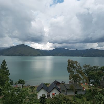The image depicts a serene natural landscape with a vast body of water, likely a lake, dominating the mid-ground. The water is a calm, turquoise hue, reflecting the overcast sky. In the distance, rolling hills covered in lush green vegetation form a picturesque backdrop, disappearing into a hazy horizon. The sky above is filled with dramatic, thick clouds, suggesting a potentially stormy or moody atmosphere.
In the foreground, a cluster of modern, minimalist buildings with dark pitched roofs are visible, nestled amongst verdant trees and shrubs. These buildings appear to be houses or accommodation, some with white walls and others with more natural-colored exteriors. One structure has a distinct orange water tank on its roof. A rusty-roofed shed is also present. The presence of these structures indicates human habitation or a resort-like setting. The greenery in the foreground is dense and varied, with trees of different heights and types. A white car is parked in the mid-ground near some of the buildings.
The overall scene evokes a sense of tranquility and natural beauty, with a hint of human presence integrated into the landscape. The location is identified as Kalalengkio, Indonesia.