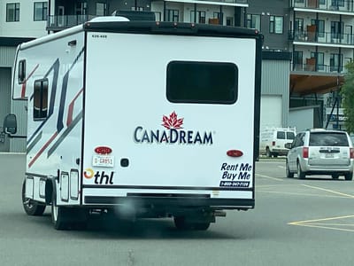 The image shows the rear of a white recreational vehicle (RV) with "CANADREAM" branding, a red maple leaf logo, and the text "Rent Me Buy Me 1-800-347-7120". The RV has a license plate that reads "O-GR851". To the right of the RV is a silver minivan with a license plate visible. In the background, there is a multi-story apartment building and what appears to be a commercial building with a large awning. The RV and vehicles are in a paved area that resembles a parking lot. Based on the visible text "Alberta", it is unlikely this scene is in Whitehorse, Canada. The license plate formatting and the word "Alberta" on the license plate strongly suggest the location is in Alberta, Canada.