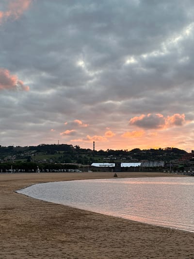 This image captures a tranquil beach scene in Gijón, Spain, likely during a sunset or sunrise, given the warm hues in the sky. The foreground is dominated by a sandy beach, with the gentle tide lapping at its edge. The water is calm, reflecting the soft colors of the sky, which is filled with dramatic, cloudy formations tinged with pink and orange.
In the mid-ground, a line of dark green trees separates the beach from a distant, rolling hill. The hill is dotted with houses and vegetation, giving it a pastoral feel. A tall, cylindrical structure with a red and white striped top, possibly a chimney or tower, stands out against the skyline on the hill. Further in the distance, along the coast, are some industrial-looking buildings, including tents or temporary structures, and what appears to be a power plant or factory with a tall chimney.
The overall atmosphere is serene and picturesque, suggesting a peaceful moment by the sea in Gijón.