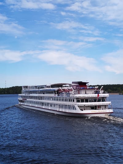 A large, white passenger cruise ship labeled "CAHKT-ПЕТЕРБУРГ" (St. Petersburg in Russian) on its stern is sailing on a body of water. The ship has multiple decks with numerous windows, a dark red and white structure on its top deck, and a small Russian flag is visible. The water is a deep blue and the ship is creating a noticeable wake. In the background, there is a strip of green trees along the shoreline and a tall, narrow antenna tower. The sky is blue with scattered white clouds, suggesting a clear day. The location is most likely the city of Myshkin, Russia.
