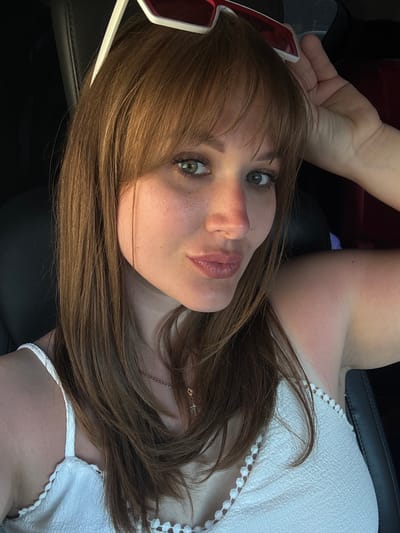 A woman with light skin and shoulder-length auburn hair, featuring bangs, is pictured in an indoor setting. She is wearing a white top with a crocheted edge and a delicate gold necklace. Red and white sunglasses are perched on her forehead. The background suggests a car interior with black leather seats. The lighting suggests it is daytime. There is no information available to identify the location as Yevpatoriya, Ukraine.