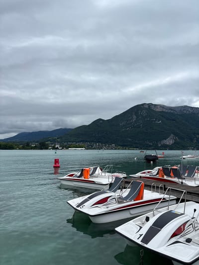 The image shows a row of pedal boats docked on a lake in Annecy, France. The lake is calm and the water is clear, with a greenish-blue hue. In the background, a range of lush green mountains rises up, their peaks partially obscured by a cloudy sky. The mountains are densely forested, with houses and buildings scattered along the foothills. 

The pedal boats are white with red and black accents. They are equipped with seats and steering wheels. Orange life vests are visible on some of the boats. The boats are tied to a wooden dock, which is partially submerged in the water. 

The sky is overcast and grey, with soft clouds that diffuse the light. The overall atmosphere is serene and peaceful. The image captures a typical scene of recreational activity on Lake Annecy, a popular tourist destination known for its natural beauty and outdoor activities.