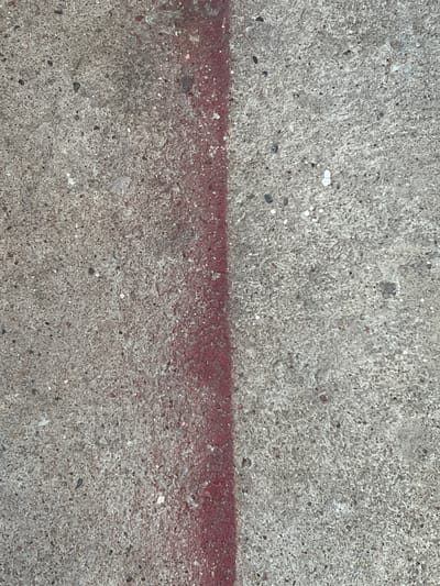 The image is a close-up, top-down view of a concrete surface with a red line painted vertically down the center. The concrete is rough and textured, with visible aggregate and small dark and white speckles. The red line is not perfectly straight and appears to be spray-painted. The background is consistent throughout the image, with no discernible features beyond the concrete and the red line. The location in the city of Ladozhskoe Ozero, Russia, is not visually discernible from the image content alone.