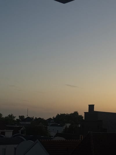The image is an outdoor photograph taken at dusk, likely from a rooftop or elevated vantage point, overlooking a residential area in Deventer, Netherlands. The sky displays a gradient of colors, transitioning from a pale blue or gray on the left to a warm, hazy orange or yellow on the right, indicating the setting sun or twilight. Silhouetted against this sky are the rooftops of houses and trees.
Several distinct architectural elements are visible. On the right side, a dark, multi-tiered roof structure with a chimney-like projection is prominent, suggesting traditional Dutch architecture. Further into the scene, other houses with tiled roofs and some white structures are discernible, interspersed with the dark green foliage of trees. A tall, slender tower, possibly a communication mast or church spire, is visible in the distance, adding to the urban landscape.
The overall mood of the image is calm and serene, characteristic of the transition from day to night. The hazy atmosphere suggests either a humid evening or possibly light pollution contributing to the diffused light. There is no visible text in the image. The focus is on the interplay of light and shadow on the urban environment and the sky.