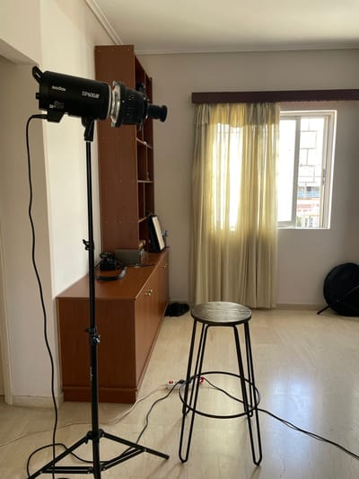 This image captures an indoor room in Vironas, Greece, set up for photography. The primary subjects include professional lighting equipment, furniture, and a window providing natural light.
In the foreground and central to the image, a black Godox DP600III studio flash unit is prominently displayed. It is mounted on a black boom arm, which in turn is supported by a light stand, extending horizontally into the room. The boom arm has "Prolux" text visible. Power cables from the flash run towards a power strip on the floor.
The room features light-colored walls and a light-toned, possibly tiled, floor. Along the back wall, a long, low brown wooden storage unit or bookshelf is visible, filled with various items. Above this unit, a window with light beige horizontal blinds allows strong natural light to stream in, casting bright patterns across the blinds and the wall. Through the upper, uncovered part of the window, residential buildings can be seen, indicating an urban or suburban environment. To the right, a minimalist stool with a black metal frame and a round wooden seat is partially visible, appearing to be stored against the wall.
The scene depicts a functional space, likely a home studio or a room temporarily converted for a photoshoot. The strong sunlight suggests it is daytime. No people are present, so there is no specific activity or event taking place, but the setup clearly indicates preparation for photography.
