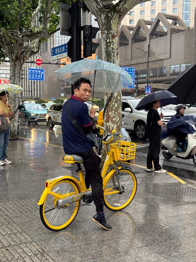 In Xuhui District, China, a man on a bright yellow bicycle navigates a wet city street in the rain. He is holding a transparent umbrella over himself and the handlebars, while another clear umbrella is held by a woman to his left. The man, wearing a dark blue polo shirt and dark pants, is looking towards the camera. The street is paved with stone tiles and reflects the overhead lights and traffic. A yellow and black sign is visible on the bicycle's frame. Several cars, including a turquoise taxi, are also on the street, their headlights illuminated. A scooter with a rider in a blue rain poncho is to the right of the man. Buildings with modern architectural designs line the background. The scene is one of everyday urban life during inclement weather.
