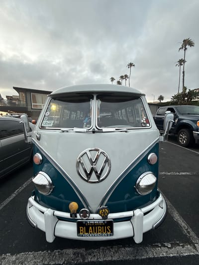 An overcast day in Oceanside, California, USA, with a weathered, two-tone Volkswagen bus, white on top and teal on the bottom, parked in a lot. The bus, featuring the iconic VW emblem on its front, has a license plate that reads "BLAUBUS" and "CALIFORNIA" and "1968". It is adorned with various stickers and a small shell decoration on the bumper. A dark-colored pickup truck is parked to the right of the bus, and a portion of a silver car is visible to the left. In the background, a contemporary building with large windows and palm trees line the street, suggesting a coastal setting.