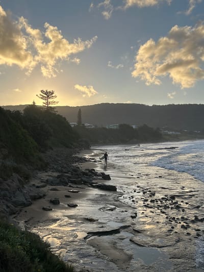 The image depicts a beach scene at dusk in Wollongong City Council, Australia. The sky is partly cloudy with golden light. In the distance, there are dark hills or mountains. The beach is sandy with some rocky areas. A person is walking with a surfboard along the shoreline. The water's edge is visible, with some waves breaking.