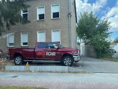 This media file shows a dark red Dodge Ram 2500 pickup truck parked on a paved area next to a building. The building appears to be a multi-story residential or commercial property with textured gray walls and white-framed windows covered by light-colored blinds. Trees and foliage surround the building and the paved area. A metal gate is visible in the background.
The truck has "GO 102" emblazoned in white on its side. The text "RAM 2500" is also visible on the truck. The truck is parked on a slight incline. A yellow marker post is positioned in front of the truck, next to a concrete border. The ground is paved with interlocking gray bricks. The scene suggests an outdoor setting during daylight hours, with a mix of vegetation and urban structures. The weather appears partly cloudy. The location is identified as Großbeeren, Germany. No people are visible in the frame.