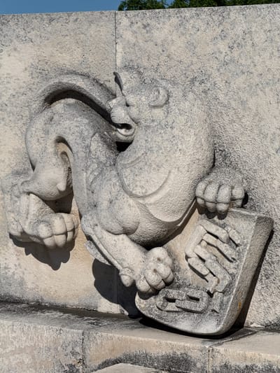 This is a close-up, eye-level, outdoor, daylight photograph of a stone carving located in Shipka, Bulgaria. The carving depicts a stylized mythological creature, possibly a griffin or a lion, with a lion's head and a scaled body. The creature is shown in profile, facing right, with its mouth open, revealing sharp teeth. It has a long, curled tail and powerful legs. The creature's right forepaw is resting on a shield or crest, which is carved with intricate geometric patterns, resembling a chain link or a stylized coat of arms. The stone appears weathered and aged, with dark patches and textures indicative of time and exposure to the elements. The background is a rough, textured stone wall, and a bright blue sky is visible above the wall. The overall scene is one of ancient craftsmanship and symbolic representation.