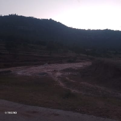 A landscape depicts a dark mountain range silhouetted against a pale sky. Terraced hillsides descend toward a foreground of rough terrain, marked by exposed soil and sparse vegetation. A pile of light-colored material sits near a dirt path. The overall scene evokes a sense of rural isolation under twilight conditions. The lower left corner displays the "TECNO" brand.