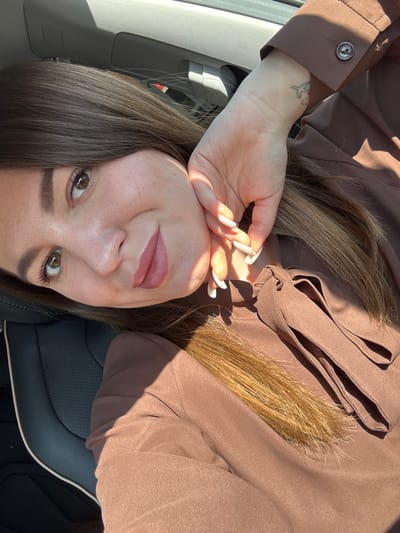 A female individual is depicted in a close-up, selfie-style photograph taken within the interior of a vehicle in Likino-Dulyovo, Russia. The subject has long, dark brown hair, brown eyes, and is wearing a brown long-sleeved garment with a tie-neck detail. Her lips are painted a reddish-brown. The left hand, adorned with long, white-tipped fingernails, is positioned near the left side of her face, partially obscuring it with shadows. A small airplane tattoo is visible on her left wrist. Strong, direct sunlight illuminates the scene, casting distinct shadows across her face and clothing. The vehicle interior features dark seating with visible stitching and a light-colored headliner in the background. The individual's gaze is directed slightly upward and to the left within the frame.
