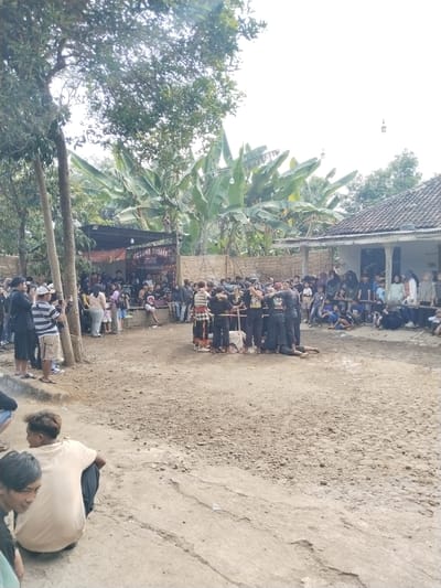 The image shows an outdoor gathering in Mojodoyong, Indonesia. A crowd surrounds a central group of people who appear to be performing or participating in an event on a dirt ground. Some individuals in the crowd are taking pictures. In the background, there is a raised stage, a house with a tiled roof, and trees including banana trees. Overhead, there are hanging lights. The ground is uneven. Several people are seated on the ground in the foreground and to the right of the frame.