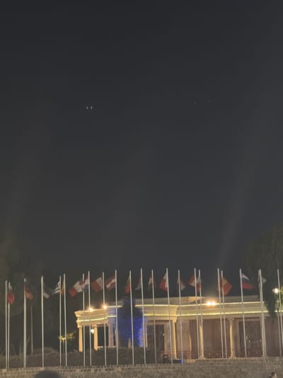 The image depicts an outdoor nighttime scene with a building illuminated by lights. A series of flags are displayed on poles extending horizontally from the building's facade, creating a decorative line. Some of the flags are visible as red, white, and yellow, suggesting they may be national or organizational flags. The building appears to have a traditional architectural style, with stone elements and multiple windows. The overall atmosphere is festive, possibly indicating an event or celebration. The sky is dark, implying it is nighttime, and the lighting from the building casts a warm glow on the surroundings. There are no visible people or text in the image. The location context is Nicosia, Cyprus.