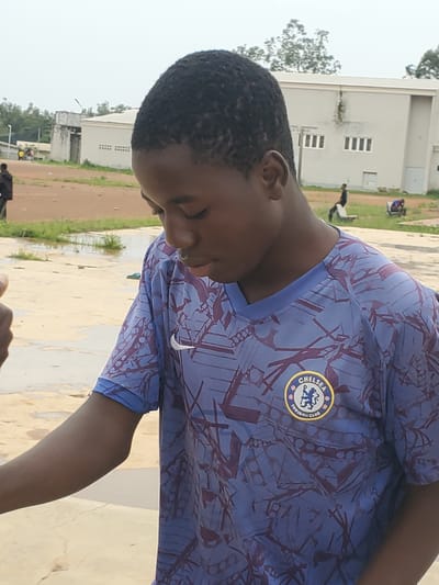 An outdoor shot captures a young Black boy wearing a blue Chelsea Football Club jersey with a purple geometric pattern and a white Nike logo. He is looking down and to his right, with his head tilted. His short dark hair is visible. In the background, there are buildings with white walls and rectangular windows, a dirt ground with puddles of water, and some green vegetation and trees. The sky is overcast and light. There are other people in the background, some standing and some sitting on benches. The overall setting appears to be a sports field or schoolyard. The scene is located in Pankshin, Nigeria.