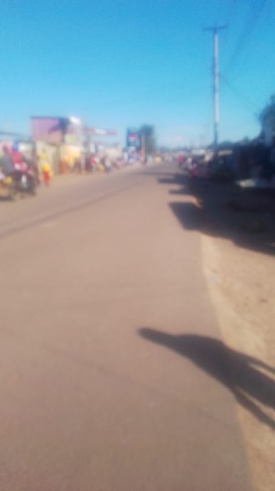 The image displays a heavily blurred, sunlit street scene in Ongata Rongai, Kenya, viewed from a low perspective looking down the length of a paved road. A light-colored asphalt road occupies the foreground and center, extending into the distant horizon under a clear, bright blue sky. On the left side of the road, multiple indistinct structures, likely commercial buildings or market stalls, are visible, characterized by soft blurs of various colors. Numerous blurred human figures are present along the left side of the road, indicating pedestrian activity. Faint outlines of what appear to be motorbikes or small vehicles are also discernible on the left. The right side of the road features an unpaved dirt shoulder, with a tall, light-colored utility pole prominent in the midground, extending vertically into the sky. Long, dark shadows from unseen objects on the right fall across the shoulder and onto the paved road. A distinct, elongated human shadow is cast on the paved road in the lower right foreground, extending towards the center of the frame, indicating the position of the photographer.
