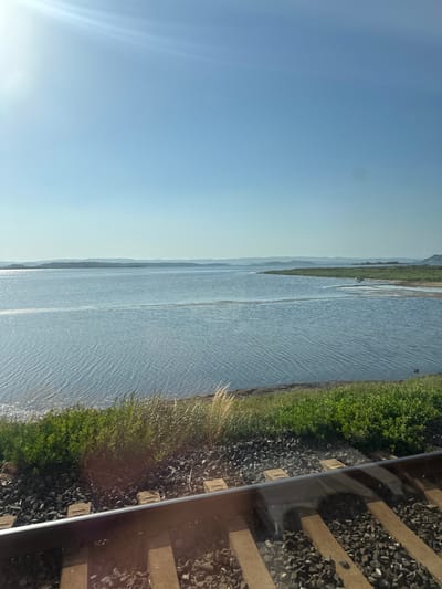 The image shows a body of water next to a railway track. The water is calm and reflects the sky, which is clear and blue with some light rays. There are green fields and hills in the distance. The railway track is made of gravel and concrete sleepers. The sun appears to be low in the sky, suggesting it might be morning or evening. The scene is peaceful and serene, with no people or animals visible. The location is Bages, France.