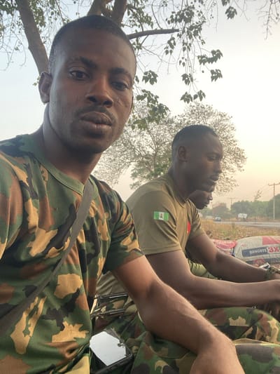 Two men in military uniforms are in the shot, with the closest man looking directly at the camera. The man in the background is wearing a tan shirt with the Nigerian flag on his left shoulder. There are multiple bags of Dangote cement in the background as well as bare trees. The sky is overcast with a light hue.