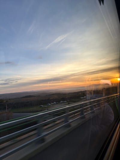 The image captures an outdoor scene at either sunrise or sunset, viewed through a glass panel, possibly from an elevated position. The orientation of the image is rotated 90 degrees clockwise, so for clarity, the description assumes a standard landscape orientation with the sky at the top.
The upper portion of the frame is dominated by a vibrant sky, transitioning from a soft, light blue on the left to intense hues of orange, yellow, and golden light on the right. Faint, wispy clouds or contrails stretch horizontally across the bluer parts of the sky. A bright, intense light source, presumably the sun, is visible on the far right edge, casting a warm glow across the scene.
Below the colorful sky, a distant, hazy landscape extends horizontally across the middle of the image. This landscape appears as dark, undulating hills or low mountains, suggesting a wide expanse of land.
The bottom section of the image reveals human-made infrastructure. Several parallel, horizontal bands, appearing as lanes or structural beams, stretch from the middle-left towards the bottom right. These bands are dark grey and green, suggestive of a multi-lane roadway, railway, or an elevated track or bridge. To the left of these lanes, there's an area of green ground, possibly grass, where several vehicles (likely cars) are parked in a row. Further back from the parked cars, a few small, boxy white buildings are visible.
Numerous reflections are present on the glass surface through which the image is captured. In the upper-right area of the sky, a reflection of a white, rectangular object with some dark text or a logo is visible, resembling a flag or banner. In the lower-right quadrant, superimposed over the infrastructure and horizon, a distinct reflection of a person's upper torso and arm is visible. The person appears to be wearing a light-colored, soft, or fluffy garment, possibly a robe or blanket. The reflections of the buildings and parked cars also appear slightly ethereal or layered with the actual view, indicating the presence of glass.
The warm, golden light and the sun low on the horizon strongly suggest the time of day is either sunrise or sunset. No specific text is clearly legible, though text is hinted at on the reflected white banner.
