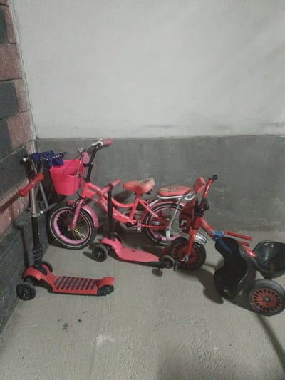 In a dimly lit, concrete-floored room, possibly a garage or storage area, a collection of children's ride-on toys are parked. The walls are a plain, off-white color, with a section of what appears to be a tiled wall in the background on the left. The floor is a rough, grey concrete surface.
Arranged in a semi-circle are two pink children's bicycles and three scooters, one silver and two pink. The larger pink bicycle has a pink basket on the handlebars. A smaller pink bicycle or tricycle with a red frame is positioned behind it. The silver scooter stands to the left, with blue handgrips. A pink scooter is in the center, and another pink scooter is to the right, partially obscured. A black trike with a wide seat and red wheels is positioned in the far right of the frame. The scene suggests a domestic or storage setting for children's recreational equipment. The specific location of Mady, Kyrgyzstan is not visually determinable from the image.