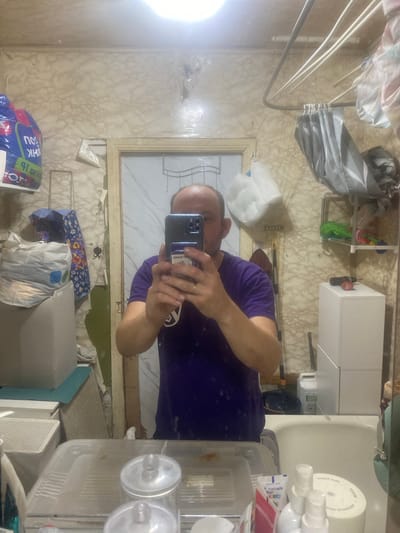 The image depicts a man taking a selfie in a bathroom. The bathroom appears to be in a state of disarray. The walls have a marble-like pattern, but they are peeling and stained in places. A clothes drying rack is mounted on the wall above the tub, with laundry hanging from it. Various items are stored on shelves and surfaces, including bags, boxes, cleaning supplies, and what appear to be personal care products. A washing machine is visible in the background. The man is wearing a purple t-shirt and has a ring on his left hand. The overall impression is one of a lived-in, functional, but somewhat neglected space. The location is described as Pereslavskoe, Russia, which suggests a typical residential bathroom in that region.