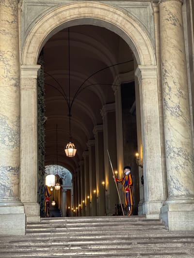 The image depicts an interior architectural scene within the Apostolic Palace, Città del Vaticano, Vatican City, characterized by a long, vaulted corridor with a visible Swiss Guard.
In the foreground, a wide, shallow flight of worn stone steps leads up to a grand, multi-layered archway constructed of light-colored stone. This archway is flanked by two large, fluted marble columns, each exhibiting prominent greyish-purple veining against a light beige background.
A Swiss Guard stands prominently on the second step from the top on the right side of the archway. He wears the distinctive red, yellow, and blue striped uniform, a black beret, and spectacles, holding a wooden halberd vertically with both hands. Another Swiss Guard, similarly uniformed, is partially visible further down the corridor on the left, also holding a halberd.
The corridor recedes into the background, revealing a vaulted ceiling with dark structural ribs. Multiple ornate lanterns, each featuring a six-sided glass shade emitting a warm glow, hang from the ceiling at varying intervals. The closest lantern is centrally positioned within the main archway. Along the right wall of the corridor, a series of wall-mounted sconces with electric candle-shaped lights cast additional warm illumination on the stone pillars and walls. The left side of the corridor appears to have a large, dark, possibly metal gate or door adorned with numerous decorative protrusions. At the far end of the corridor, another archway leads to a brightly lit area, revealing intricate architectural details, possibly including statuary or relief work. The overall lighting is dim, emphasizing the illuminated fixtures and the figures of the guards.