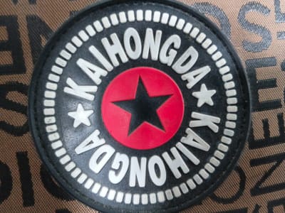 The image shows a close-up, eye-level shot of a circular patch with white lettering on a black background, set against a brown patterned fabric. The patch features the words "KAIHONGDA" arranged in a circle, with the letters embossed and white. Within the circle of text, there is a red circle containing a black five-pointed star. The surrounding fabric has a repeating pattern of dark letters, possibly a logo, on a lighter brown base. The lighting appears to be natural and even, with no harsh shadows. The scene is static and focused on the patch and its immediate surroundings. There are no people, actions, or interactions visible. The provided information about the location in "Tecnimont camp, Russia" is not visually supported by the image content.