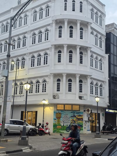 This is a vertical-format, eye-level shot of a white, multi-story building with numerous windows. The building's facade features architectural details like columns and decorative elements above some of the windows. The windows themselves are large and rectangular, with some arched tops. A street runs in front of the building, with a sidewalk.
Several objects are visible on the street and sidewalk. A white car is parked on the right side of the frame, and two motorcycles are parked behind it. A person is riding a red and white motorcycle in the foreground, partially obscuring the building. Streetlights are visible, one of which is illuminated, suggesting it might be late afternoon or evening. A mural is painted on the lower part of the building's wall, depicting several buildings.
Text visible in the image includes "HERITAGE MEDAN," "Heritage Boutique HOTEL MEDAN," "BY TONGA," "BREW BEAN COFFEE SHOP," and "PT. INDUSTRI." The presence of "HOTEL MEDAN" suggests the building may be a hotel. The overall scene appears to be in a city, likely during daylight hours, though the illuminated streetlight suggests it could be nearing dusk or early morning. The weather appears to be overcast.