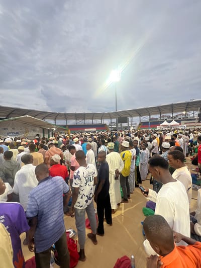A wide-angle, slightly elevated perspective captures a large-scale outdoor gathering of thousands of predominantly male individuals on the field of a stadium in Zanzibar City, Tanzania, consistent with Amaan Stadium. The sky is overcast, with a bright floodlight on a tall pole casting a significant flare from the right side of the image. The stadium's multi-colored seating (red, blue, green) is visible in the background, mostly empty. The ground where the crowd stands is covered with a light-colored mat or tarp. Many attendees wear kufis (prayer caps) in various patterns and colors, along with a mix of traditional white robes and casual clothing. White event tents are erected on the left side, one featuring text including "JBC" and "IFTAR KITA." Individuals in the foreground are standing, some looking at mobile phones. Red and green bags or prayer mats are distributed on the ground. The scene depicts a densely packed assembly, indicating a significant community or national event, likely an Iftar gathering.