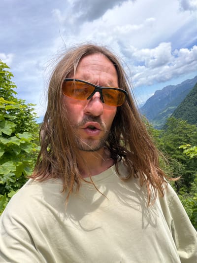 An outdoor, eye-level portrait features a male individual, located in Љевишта, Montenegro. The subject has long, light brown hair, which appears disheveled, potentially by wind. He wears black-framed, orange-tinted wrap-around sunglasses. His mouth is open in a slightly O-shaped, pursed expression, and he exhibits light facial stubble. He is clad in a plain, loose-fitting, light olive green crew-neck t-shirt. The background displays a natural landscape with dense green foliage, including trees and bushes, on both sides of the subject. Distant mountains, covered in vegetation, are visible beneath a partly cloudy sky, indicating a sunny outdoor setting.