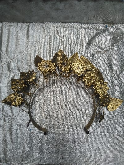 A metallic, gold-colored ceremonial headband, featuring an array of stylized leaf and eight-petaled flower ornaments, is positioned on a silver-colored, highly textured fabric surface. Each flower ornament includes a central boss surrounded by small spherical embellishments. The headband's base consists of a thin, dark, curved band, showing signs of patina. The silver fabric displays consistent horizontal and diagonal creasing, laid beneath a darker, undefined surface visible along the top edge of the frame. This scene is located in Тохта, Kazakhstan.