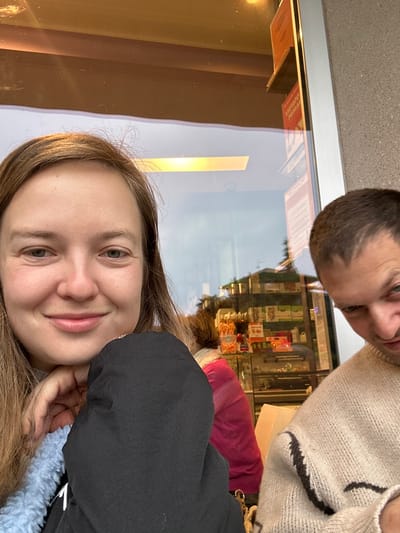 The image shows two individuals indoors, positioned in front of a large window at a location in Longone al Segrino, Italy. A female subject occupies the left side of the frame, smiling directly at the camera. She has light brown hair, fair skin, and light-colored eyes. Her right hand is placed under her chin. She is wearing a light blue fuzzy garment over a dark, possibly black, outer layer. To her right, a male subject is partially visible, cropped from the mid-face downwards. He wears a light beige knit sweater with a dark, abstract, wave-like pattern. His gaze is directed downwards and to the left, exhibiting a slight smirk.

The background is dominated by the reflective window, which displays elements from both the interior and exterior environments. Reflected within the establishment are shelves stocked with packaged food items, a display case containing various goods, and a linear overhead light fixture. A partial reflection of a third individual, with reddish-brown hair and wearing a pink top, is discernible deeper within the reflected interior space. Exterior reflections indicate foliage or trees and architectural structures. Two red signs are visible as reflections on the window; one clearly reads "VIETATO FUMARE" (No Smoking) in white text on a red background. A portion of the building's exterior wall, light grey and textured, is also visible adjacent to the window.