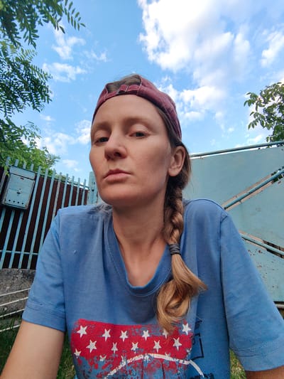 A woman with blonde hair braided into a plait, wearing a red baseball cap and a blue t-shirt with a patriotic design, is captured in a close-up, low-angle selfie. She is looking directly at the camera with a neutral expression. Behind her, a weathered, light blue metal gate stands partially open, with a rusty mailbox attached to it. The background consists of a clear blue sky with scattered white clouds, and green foliage from trees framing the top left and right of the image. The scene appears to be outdoors, possibly in a rural or suburban setting, given the fence and mailbox. The location is identified as Ароматновское сельское поселение, Ukraine.