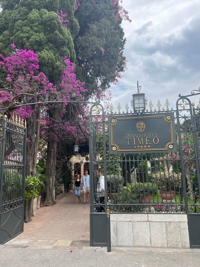 This is a vertical, eye-level medium shot of a grand entrance gate to the Grand Hotel Timeo in Taormina, Italy. The gate is made of dark metal with ornate gold detailing, featuring a prominent sign that reads "GRAND HOTEL TIMEO" with five gold stars and a circular emblem above it. The gate is partially open, revealing a pathway lined with brick paving stones and lush greenery.

In the foreground, a man and a woman stand on the pathway, facing away from the camera. They are casually dressed, suggesting a warm day. Behind them, through the gate, a glimpse of the hotel's architecture is visible, along with more trees and flowering plants, including vibrant pink bougainvillea cascading over the gate and trees. A street lamp is affixed to the gate. The sky above is cloudy and overcast, indicating a potentially humid or changeable weather condition. The overall scene conveys a sense of elegance and an inviting atmosphere, typical of a luxury hotel entrance in a Mediterranean setting.