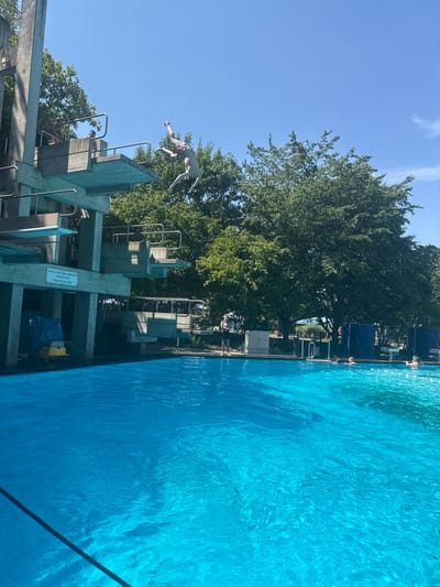 The image depicts an outdoor swimming pool on a sunny day. A person is captured mid-air, performing a dive from a high diving board. The diving platform consists of multiple levels, constructed from concrete with metal railings. Trees with lush green foliage surround the pool area, and a clear blue sky dominates the upper portion of the frame. Other individuals are visible near the diving boards and in the pool, some swimming and others relaxing. A sign on the diving structure reads "Nicht in den Sprungbereich schwimmen. Wassertiefe 3,80m," indicating a prohibition against swimming in the diving area and a water depth of 3.80 meters. The overall scene suggests a recreational setting at a public swimming facility. The presence of the inscription in German, along with the architectural style of the diving platform, suggests this location could be the Freizeitbad des Oberlinhauses in Rheinbach, Germany, which features a similar concrete diving tower and an outdoor pool.