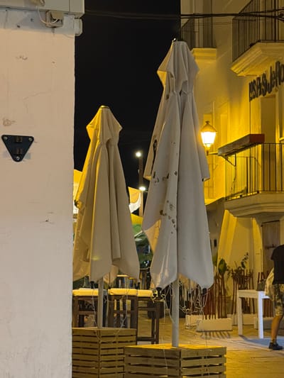 The image shows a nighttime scene in Ibiza, Spain, likely in a town or city area with outdoor seating. In the foreground, a white wall is on the left. Two large, closed, light-colored umbrellas are prominent in the center of the frame. They are positioned above wooden crates, which appear to be used as bases for outdoor furniture. Behind the umbrellas, there are tables and chairs, suggesting a restaurant or cafe setting. A yellow street lamp is visible, illuminating the scene with a warm glow. In the background, a building with a balcony and railing is lit by yellow lights. Part of a person wearing patterned shorts and a black t-shirt is visible on the far right, walking away from the camera. The overall atmosphere is relaxed and typical of an evening in a tourist destination.