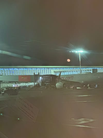 This rotated night image captures an airport scene at Nouaceur, Morocco, featuring a parked passenger aircraft of Pegasus Airlines. The airplane, prominently displaying "pegasus.com" on its fuselage and registration "TC-NCY" near the tail, is illuminated by numerous bright artificial lights from the adjacent terminal or jet bridge.

The scene is dominated by the dark night sky, with a striking red-orange circular object, likely the moon, visible above and to the left of the aircraft. Bright white and blue lights line the terminal structure, creating significant lens flare and light streaks, suggesting the photo was taken through a window. Various ground support equipment, including what appears to be a mobile stairway or jet bridge and other service vehicles, are positioned around the aircraft, indicating ongoing ground operations such as boarding, deboarding, or pre-flight preparation.

A red banner with white text, partially readable as "International Airport," is visible below the terminal structure. Further in the distance to the right, an indistinct sign with Arabic writing can be faintly discerned. The overall impression is that of a busy airport environment under the cover of night, with the red-orange moon adding a distinctive atmospheric touch.